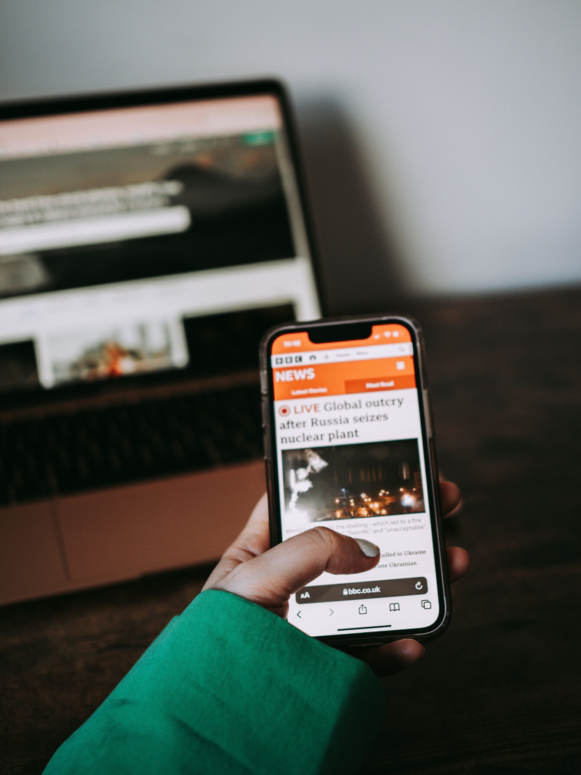 Person using smartphone for reading news online with laptop in background.